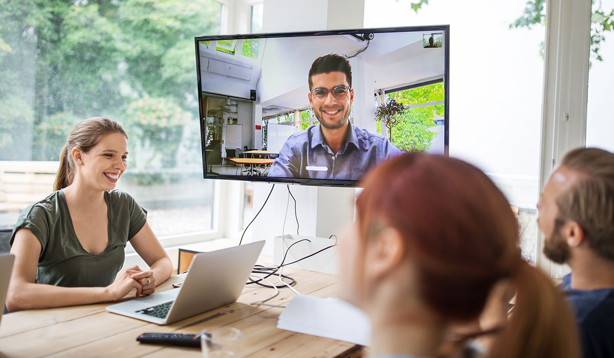 Co-workers sit around a table on a video call. There is a large TV screen showing a person on the video call. Image credit: iStock