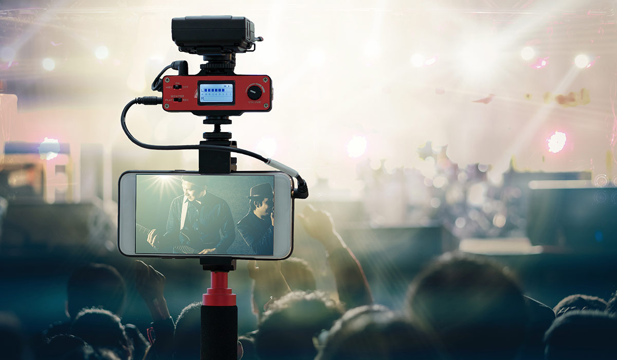 A tripod holding up an iPhone that is recording a live music event with crowds in the audience. Image credit: Adobe Stock