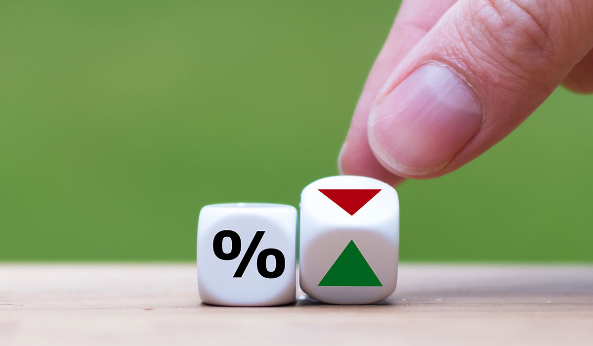 A hand rolling two dices against a green background. 1 has a percentage sign on it, and the other shows a green and red arrow. Image credit: Adobe Stock