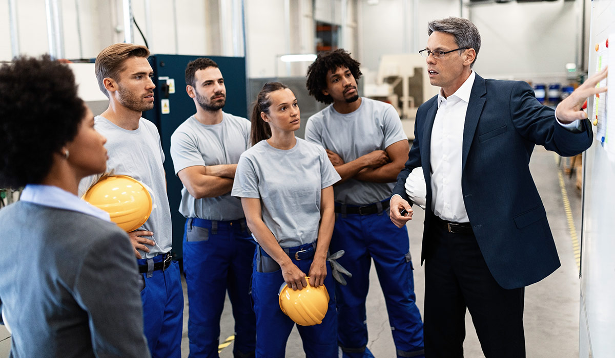 A manager in a factory points to the whiteboard as he has a meeting with his staff. They are wearing overalls and holding hard hats. Image credit: Adobe Stock