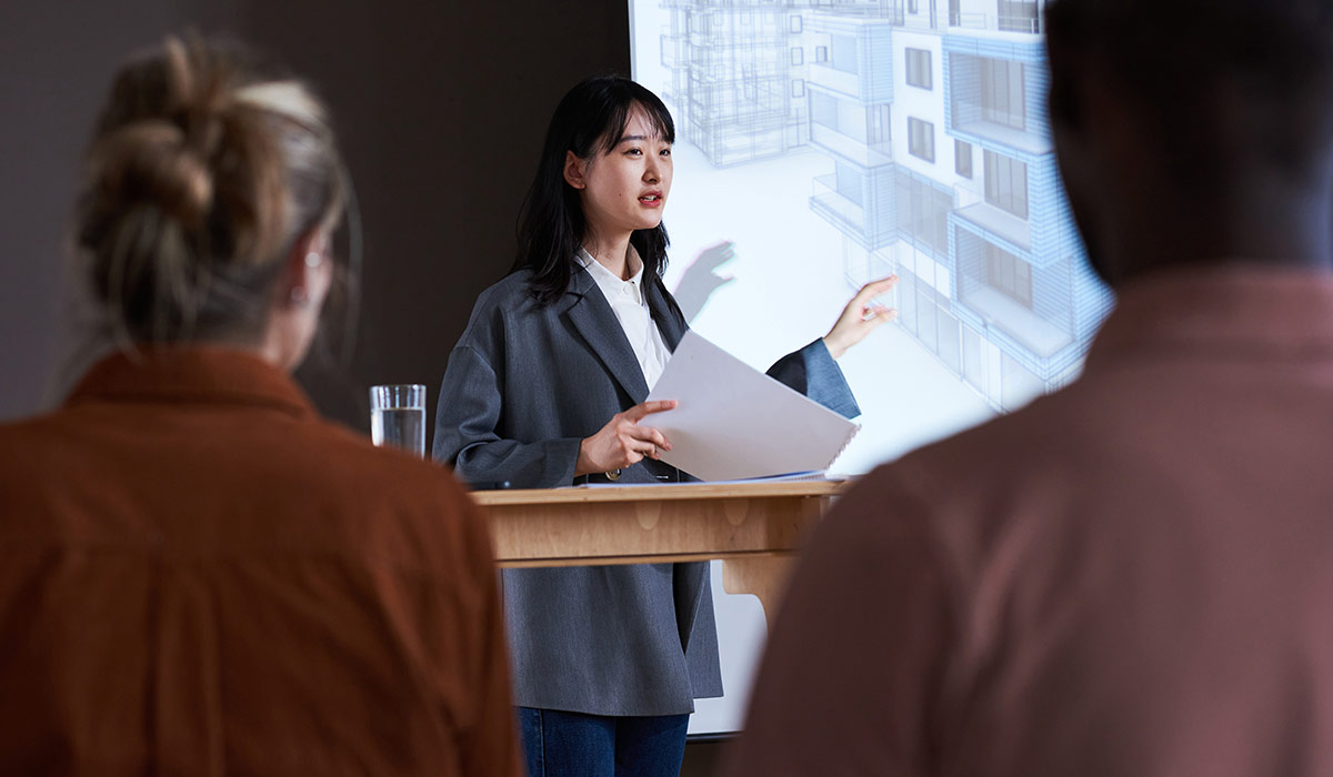 A female employee gives a presentation in front of co-workers. She is pointing to a drawing on a projector screen. Image credit: iStock